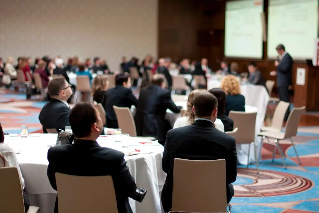 People sitting at tables during a trade show conference presentation.