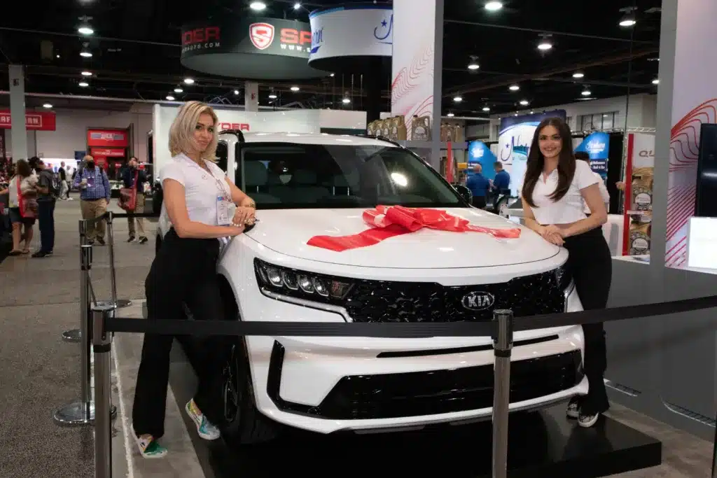 Two women standing next to a white car at a trade show.