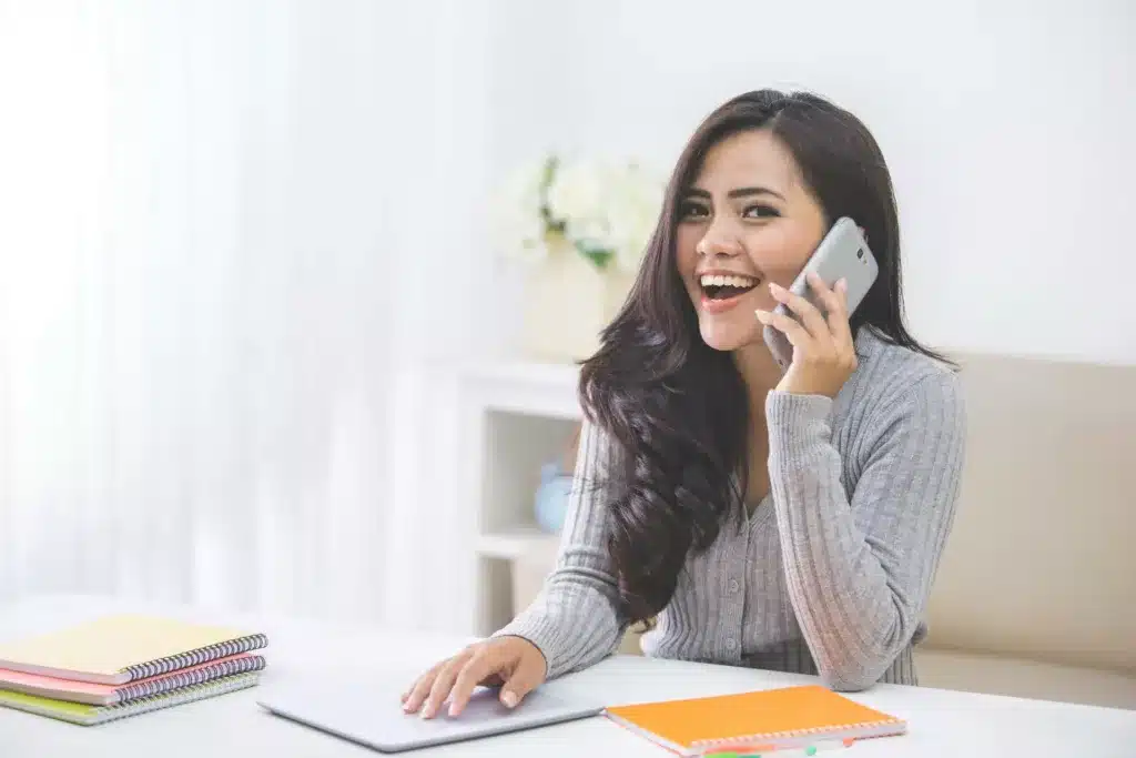 Women at a desk with a pen in her hand while she uses a cell phone to call someone.