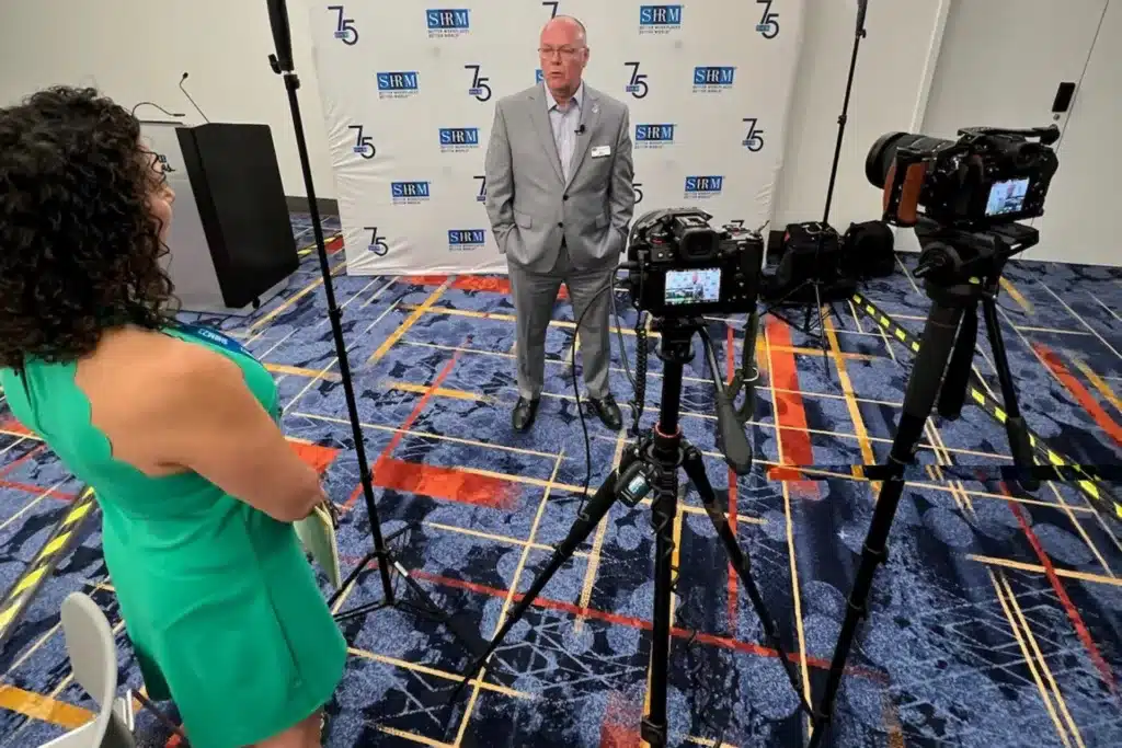 photo of a man in a gray suit talking in front of two video cameras while a woman in a green dress watches.