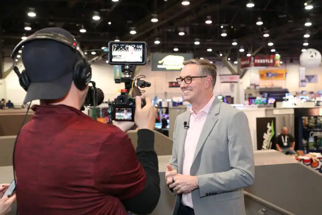 a photo of a man in a gray suit smiling in front of a video camera that another person is holding.